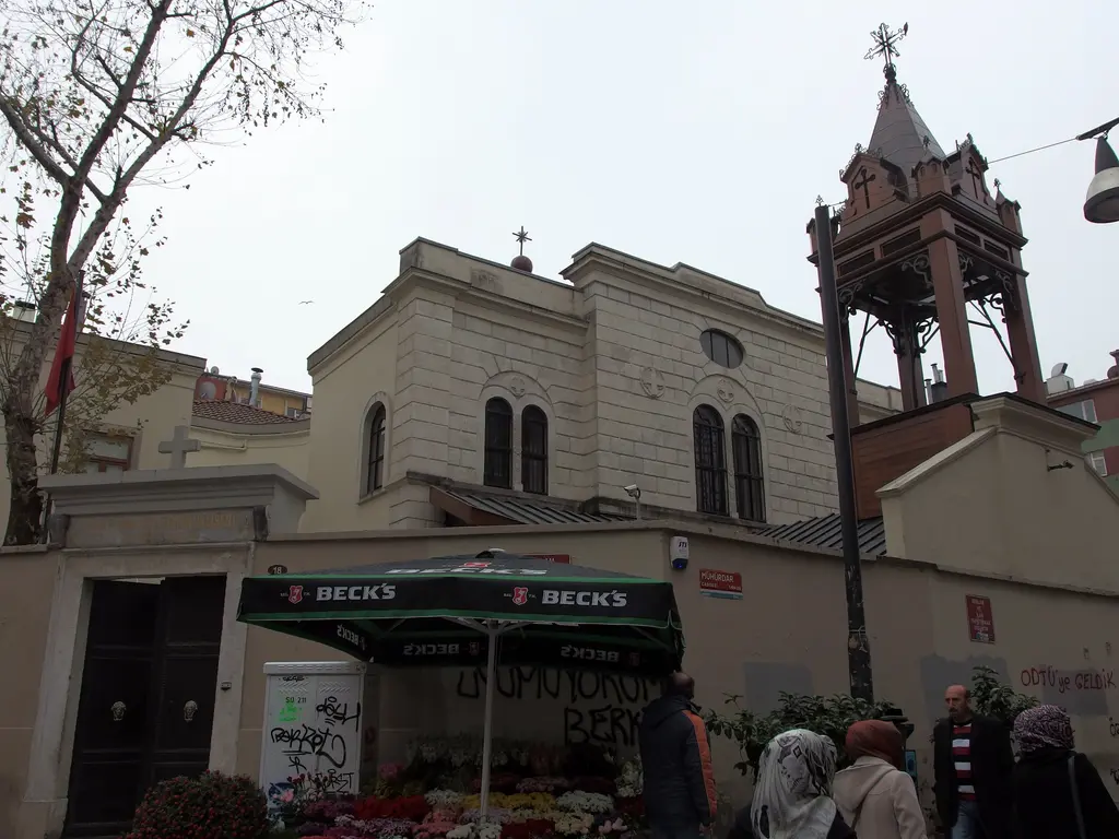 Kadıköy Çarşısı'nda Surp Takavor Kilisesi; avlu duvarı, önünde şemsiyeli büfe, çan kulesi ve yürüyen insanlar.
