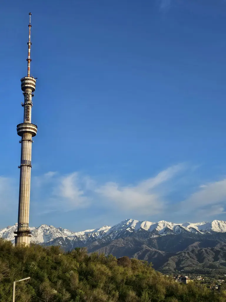 Almatı'da Kok-Töbe tepesindeki televizyon kulesi; çelik gövdesi ve tepesindeki anten, yamacı kaplayan çalılar ve arkada karlı dağ zirveleri.