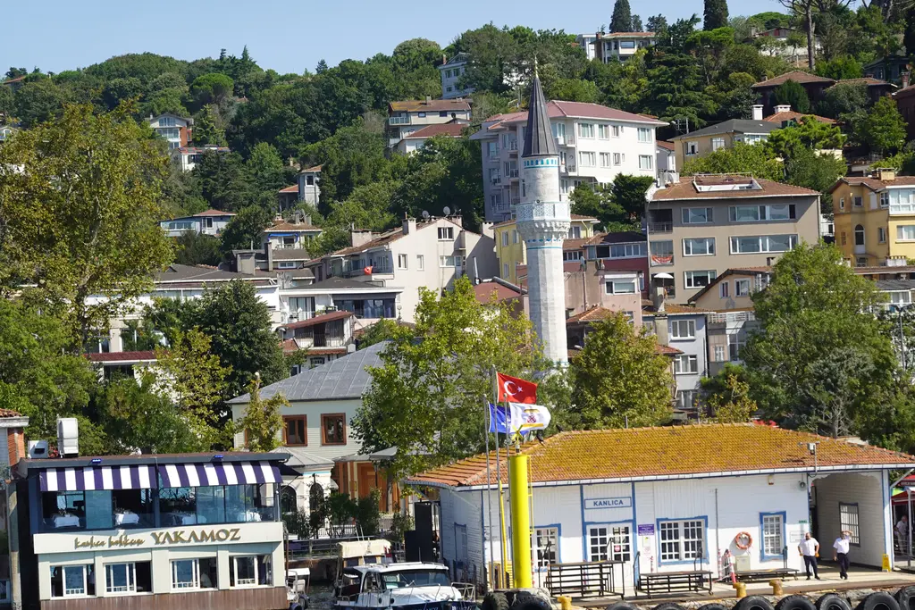 Kanlıca İskelesi ve arkası; mavi beyaz iskele binası, bayraklar, kıyıda lokanta ve yamaçta minare ile evler.