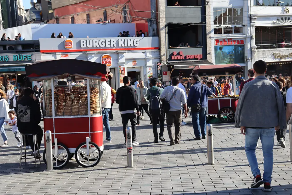 Taksim'de kırmızı bir simit arabası; tezgâhta dizili simitler, arkasında dükkân tabelaları ve kaldırımda yürüyen kalabalık.