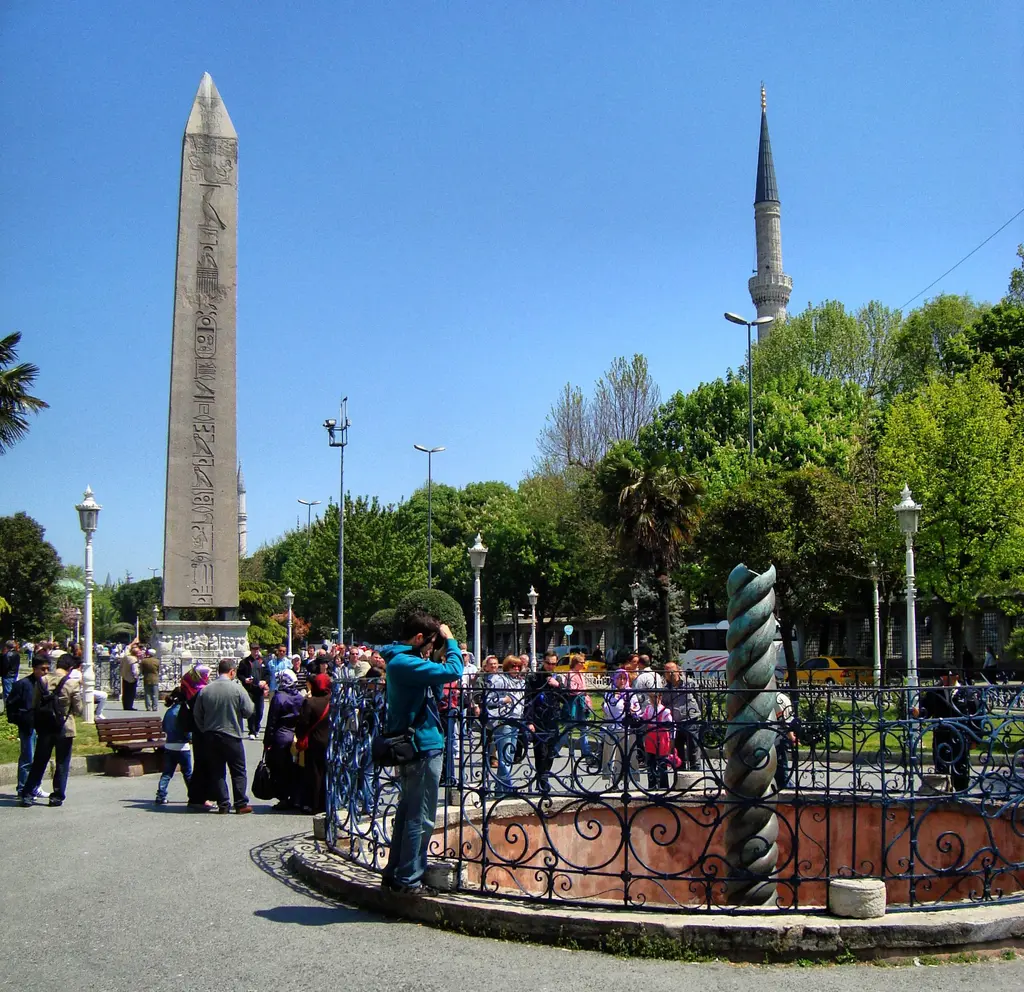 Sultanahmet Meydanı'nda Yılanlı Sütun ve arkasında Dikilitaş; parmaklık çevresinde toplanmış ziyaretçiler, ağaçlar ve bir minare.
