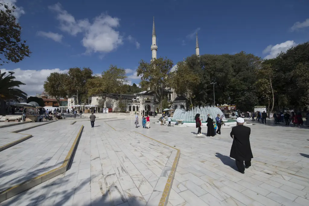 Eyüp Sultan meydanı; geniş taş döşeli alan, yürüyen insanlar, ağaçlar ve arkada yükselen cami minaresi.