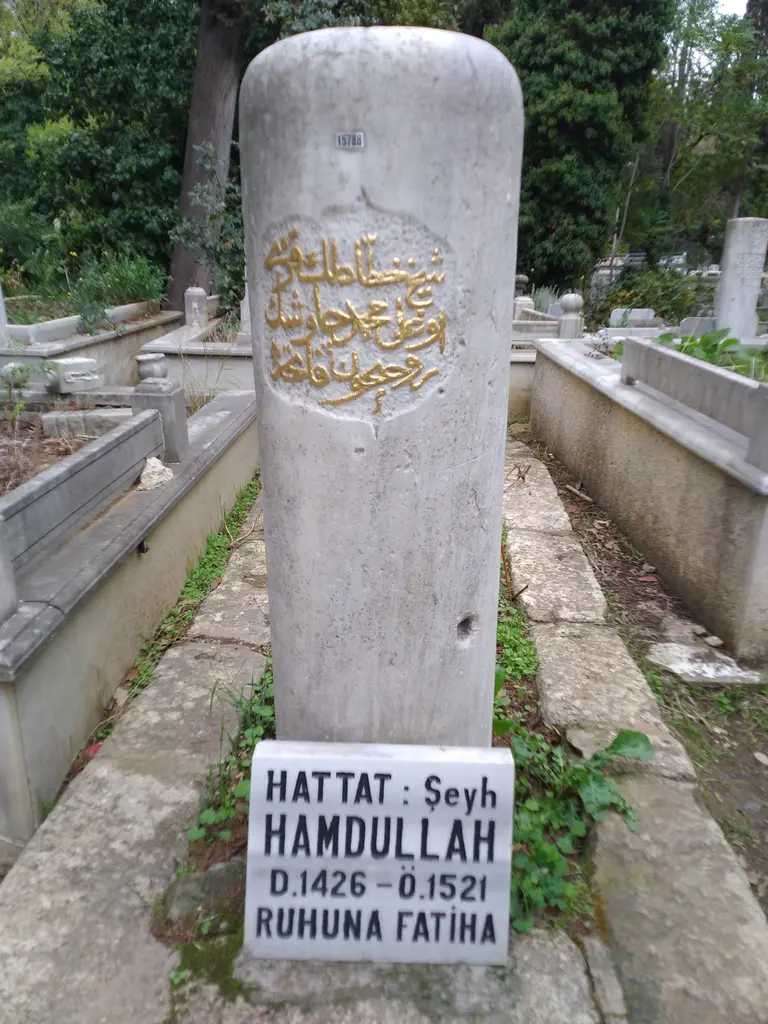 Şeyh Hamdullah'ın mezar taşı; yakından beyaz mermer gövde, altın yaldızlı Arapça yazı ve önünde "Hattat Şeyh Hamdullah" levhası.
