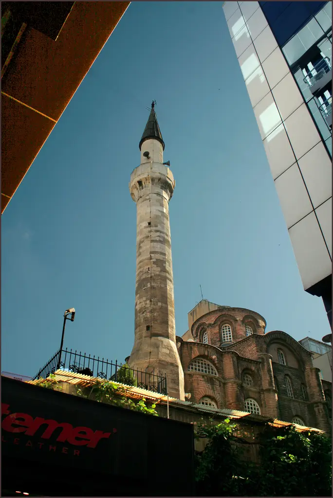 Bodrum Camii'nin minaresi sokaktan dik alttan; kesme taş gövde, şerefe, hoparlörler, koyu konik külah, sağda tuğla gövde ile payandalar, solda dükkân tabelaları.