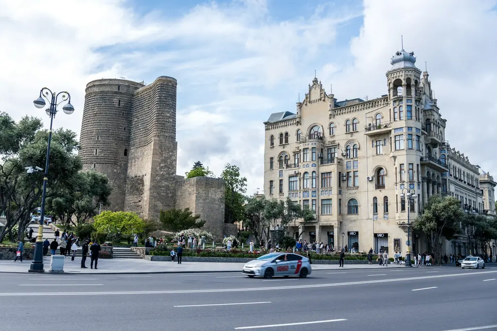 Bakü'de Kız Kalesi ile yanındaki süslü art nouveau tarzı tarihî binanın cadde seviyesinden görünümü; önde geniş bir bulvar ve geçen araçlar.