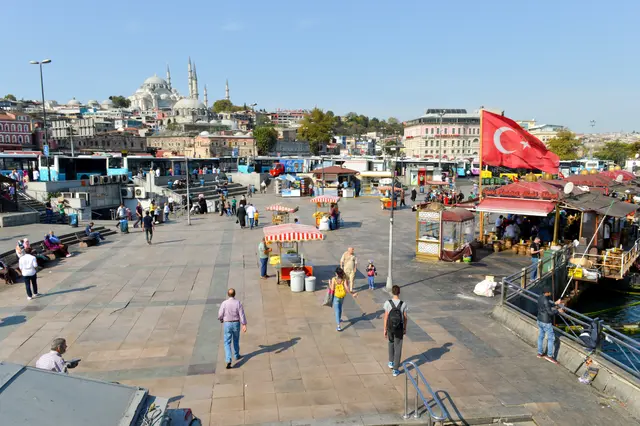 Eminönü meydanı ve kıyısı; geniş taş meydan, seyyar simitçi arabaları, balık ekmek tekneleri, Türk bayrağı ve tepede Süleymaniye Camii.