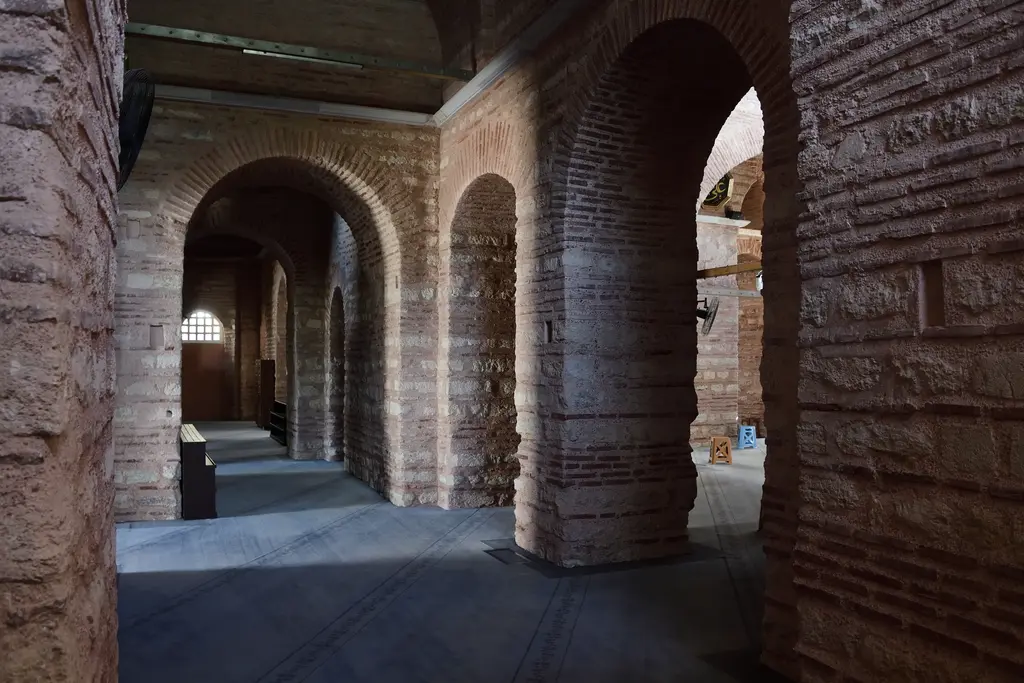 Fenari İsa Camii'nin narteksinde derin perspektif; kalın tuğla ve taş payeler, aralarındaki kemerler, gri şap zemin ve uçta aydınlık bir kemerli geçit.