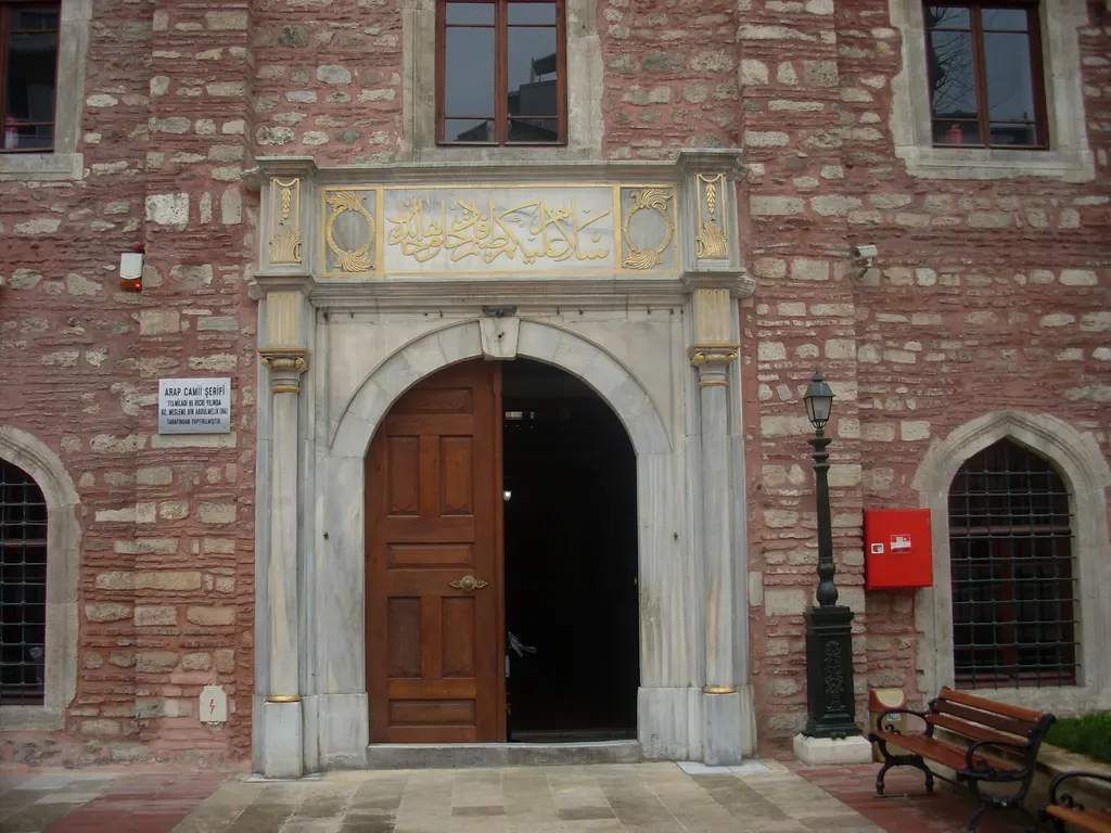 Arap Camii'nin kapısı; mermer çerçeveli yuvarlak kemerli ahşap kapı, üstünde yazı kuşağı, iki yanda pencereler ve önde bank.