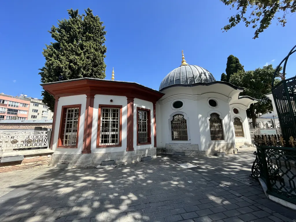 Koca Mustafa Paşa Külliyesi'nde yan yana iki türbe; solda kırmızı beyaz boyalı, demir kafesli cumba pencereli yapı, sağda kurşun kubbeli beyaz türbe, önde işlemeli döküm korkuluk.