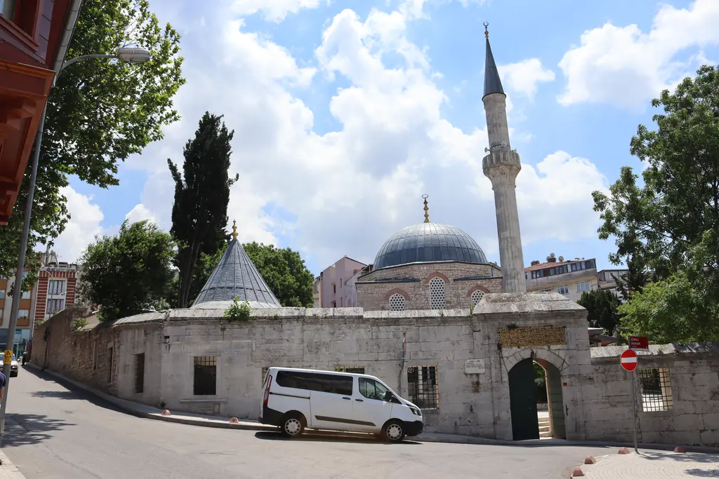 Çinili Camii'nin avlu kapısı; taş duvarda kemerli giriş, önünde beyaz bir minibüs, arkada kubbe ve minare.