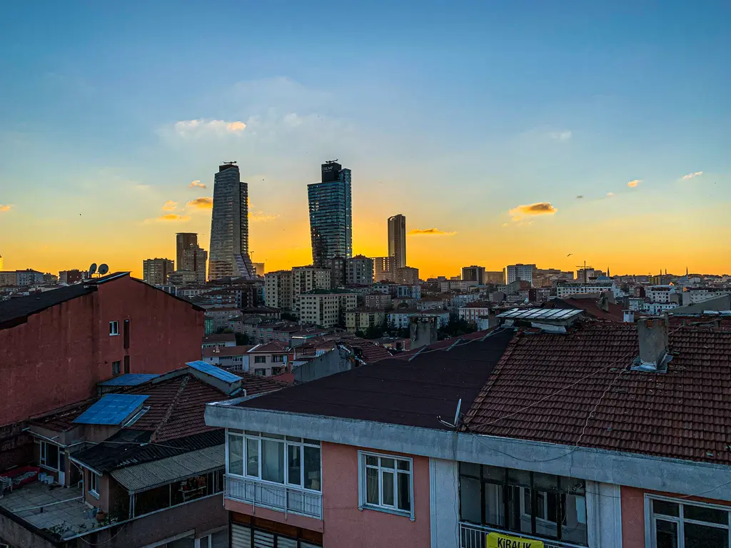 Şişli'de gün batımında Mecidiyeköy silueti; ufukta yükselen gökdelenler, önde kiremit çatılı eski apartmanlar ve turuncu mavi gökyüzü.