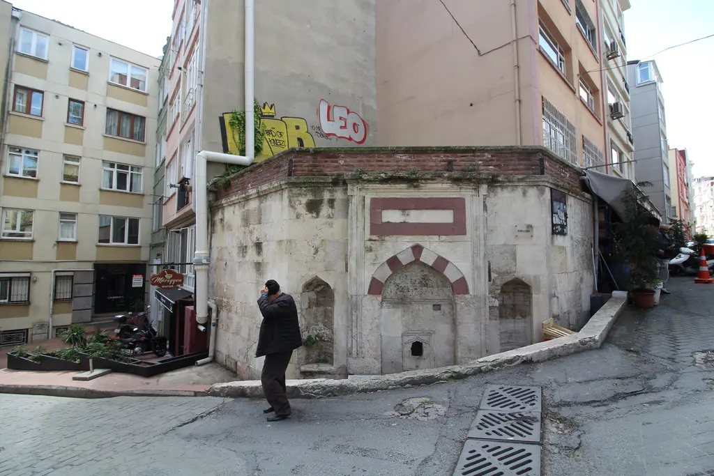 Susam Sokağı'nda köşedeki taş çeşme; kemerli nişler, yanında yürüyen bir adam ve arkada apartmanlar.