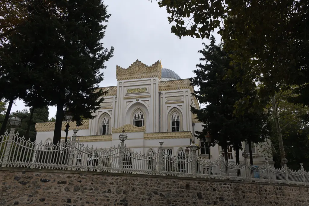 Yıldız Hamidiye Camii'nin avlusu sokaktan; taş istinat duvarı, beyaz demir parmaklık, ağaçlar ve arkada minare.