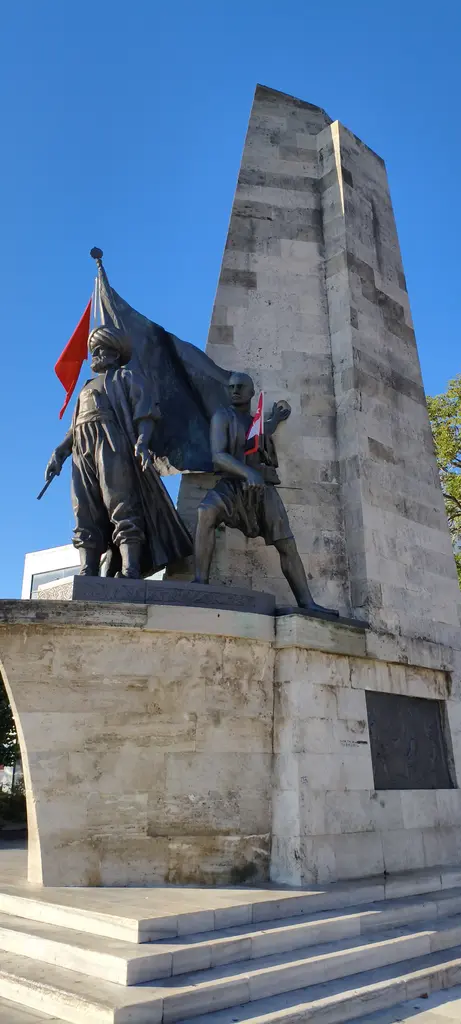 Barbaros Anıtı; mavi gökyüzü önünde bronz figürler, birinin elinde dalgalanan sancak ve altta taş kaide.